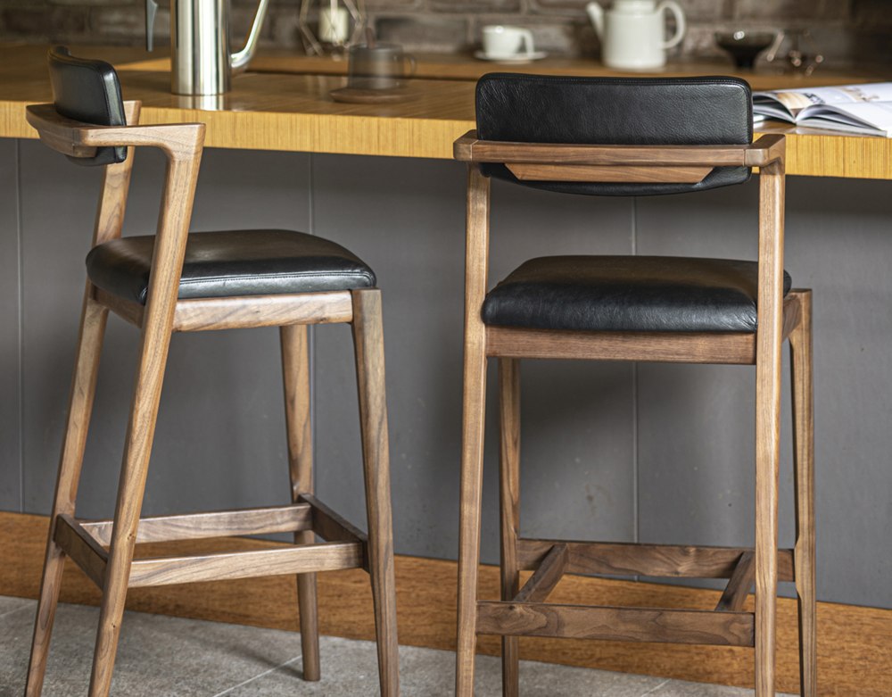 Manufacturer photograph of the Koti counter chair, showing its upholstered back, wooden sidearms and footrest